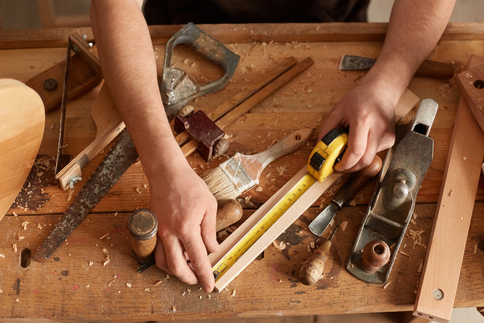 Closeup portrait of faceless Joiner carpenter working in his, workplace, different tools on wooden table, lot of equipments and wood, craftsman measuring wood block with tape in carpentry workshop.